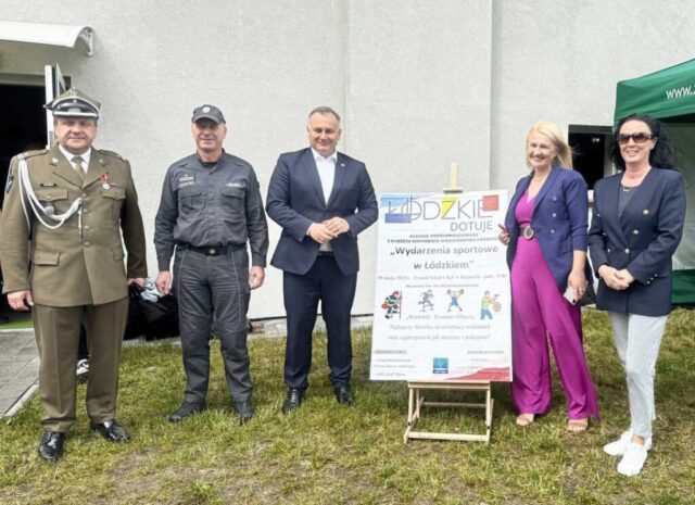 Pani dyrektor Szkoły, Pani Prezes Luks Gryf, Wicemarszałek Województwa Łódzkiego oraz przedstawiciele służb mundurowych przed budynkiem strzelnicy. Pośrodku plakat reklamujący wydarzenia sportowe w Łódzkiem i fragment namiotu firmowego Szkoły.