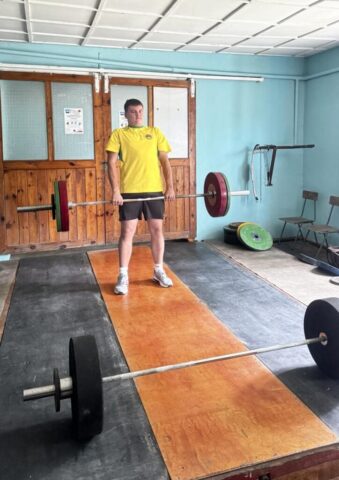 Uczestnik wieloboju sportowego w trakcie podnoszenia ciężarów wewnątrz klubu sportowego.