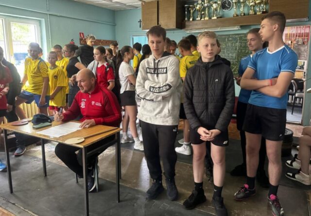 Uczestnicy wieloboju sportowego wewnątrz klubu sportowego. Przy stoliku organizator zmagań, który dokonuje zapisów w dokumentacji zawodów.