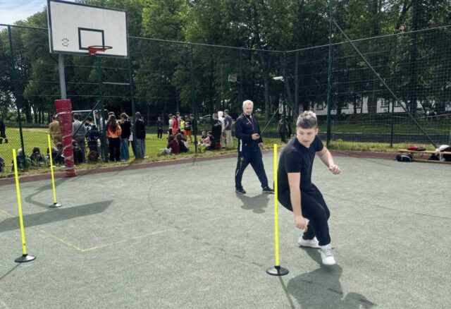 boisko orlik. Uczestnik wieloboju w trakcie biegu slalomu, w tle nauczyciel WF, trzymający stoper i obserwujący zawodnika. Widoczni też inni uczestnicy wydarzenia, obserwujący kolejne zmagania.