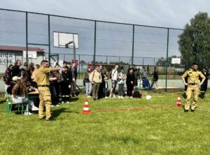 Uczestnicy wieloboju wraz z przedstawicielami służb mundurowych na zewnątrz, na terenie szkoły w pobliżu toru przeszkód. W tle boisko orlik. 