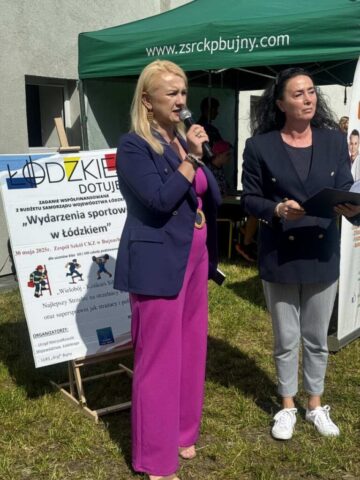 Pani Dyrektor Szkoły wraz z Panią Prezes Luks Gryf. Ceremonia otwarcia wydarzenia sportowego. Pani Dyrektor przemawia, trzymając w ręku mikrofon. W tle plakat reklamujący wydarzenie, oraz namiot firmowy i stoisko ZS CKZ Bujny.