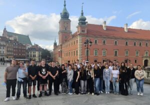 Grupa młodzieży wraz z opiekunami pozuje do zdjęcia na tle Zamku Królewskiego w Warszawie. W tle zabudowania Starego Miasta. 