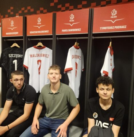 3 uczestników wycieczki w szatni kadry narodowej na Stadionie Narodowej. Widoczne także wiszące koszulki zawodników kadry piłki nożnej.