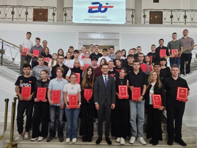 Uczestnicy i opiekunowie młodzieży w Sejmie. Pozują do zdjęcia z posłem Adrianem Witczakiem na schodach. W ręku każdy trzyma egzemplarz Konstytucji Polski.