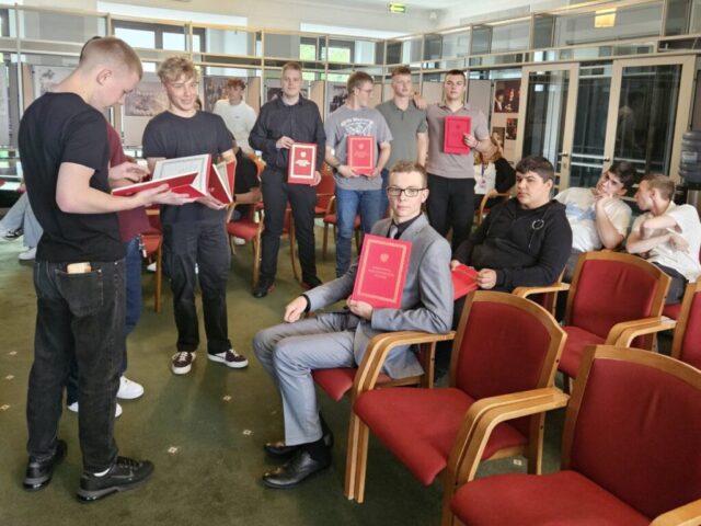 Uczestnicy wycieczki do Warszawy w gmachu parlamentu. Część z nich siedzi na czerwonych krzesłach, część stoi. W tle widoczne obrazy prezentujące życie parlamentu.
