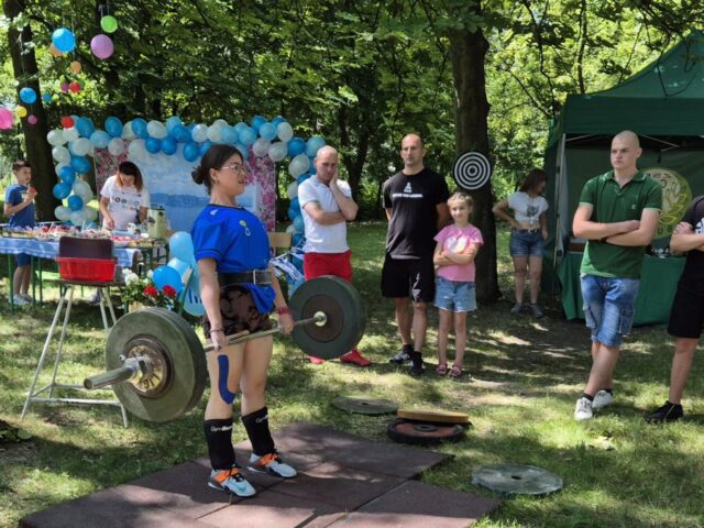 Zawodniczka w stroju sportowym podnosi ciezary. W tle widoczne stoisko technikum leśnego oraz stoisku promującego event Grecki. Widoczni także widzowie obserwujący konkurencję podnoszenia ciężarów.