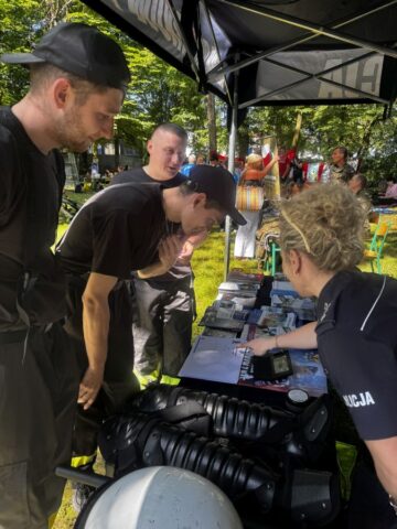 Stoisko Policji, uczestnicy festynu zawodowe lato w Bujnach słuchają objaśnień pani policjantki. Na stoisku ulotki oraz wyposażenie policyjne. W tle inne stoiska.