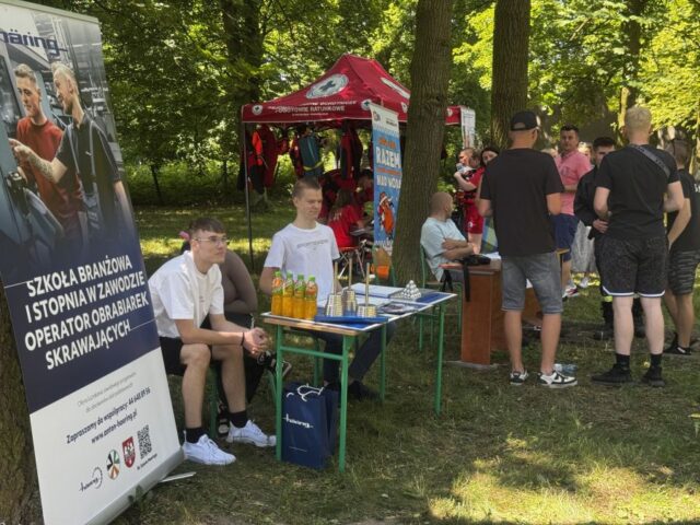 Na pierwszym planie po lewej stronie stoisko Szkoły branżowej wraz z uczniami siedzącymi przy ławce. W tle stoisko WOPR oraz uczestnicy festynu Zawodowe Lato w Bujnach.