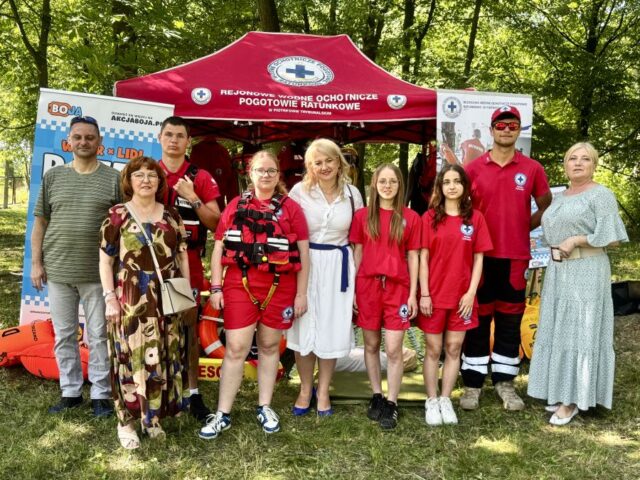 Młodzież- członkowie WOPR w czerwonych strojach z logo WOPR, Dyrekcja, opiekun szkolnego WOPR wraz z przedstawicielem ratownictwa medycznego na tle stoiska WOPR. Po bokach stoiska widoczne rollupy.