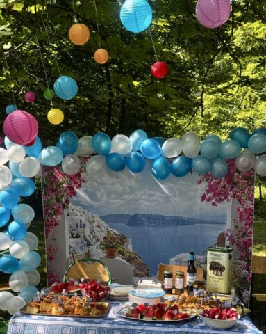 Stoisko Event Grecki. Ozdoby z balonów białych i niebieskich oraz kolorowe kule papierowe. Na materiale zdjęcie przedstawiające typowy krajobraz grecki. ławka przykryta obrusem koloru białego i granatowego. Na ławce owoce, ciasta, puszka oliwy z oliwek oraz oliwki w słoikach.