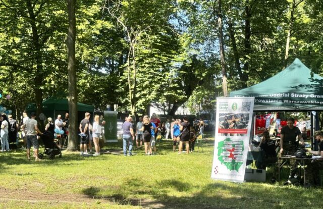 Stoiska promocyjne podczas festynu Zawodowe Lato w Bujnach. Na pierwszym planie po prawej stronie stoisko Oddziału Straży Granicznej. W tle stoisko Technikum Leśnego, uczestnicy i organizatorzy festynu Zawodowe Lato w Bujnach.