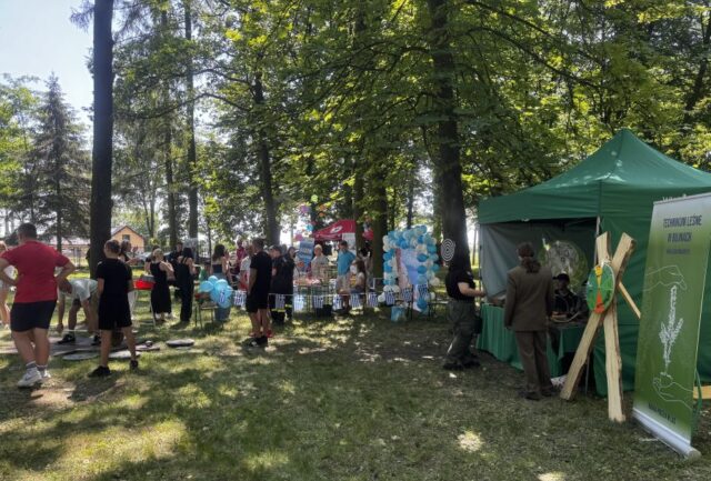 Stoisko technikum leśnego z uczniami, jeden uczeń w mundurze. Widoczny rollup Technikum, leśne koło fortuny oraz zawieona tarcza do gry w darta. W tle stoisko event Grecki, WOPR, uczestnicy i organizatorzy festynu Zawodowe Lato w Bujnach.