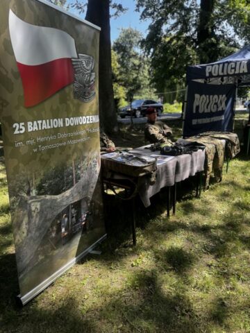 Stoisko 25 Batalionu Dowodzenia, po lewej rollup promocyjny. Ławki, na których są ubrania moro oraz sprzęt wojskowy, żołnierze siedzący przy ławkach. W tle widoczny namiot promocyjny policji.