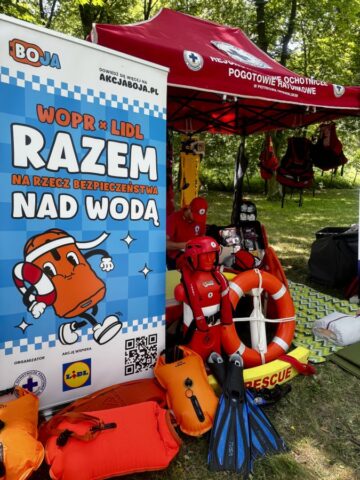 Stoisko Wodnego Ochotniczego pogotowia Ratunkowego, namiot koloru czerwonego, rollup reklamujący bezpieczeństwo nad wodą. Widoczny także profesjonalny sprzęt WOPR.