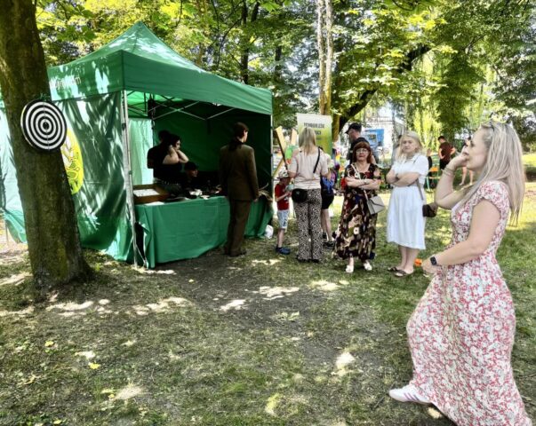 Stoisko leśne podczas Zawodowego Lata w Bujnach, uczniówie obsługujący stoisko, jeden w uczniów w umundurowaniu Technikum Leśnego. Zielony namiot promocyjny, na pierwszym planie kobieta gra w rzutki. W tle dyrekcja Szkoły oraz inni uczestnicy wydarzenia.
