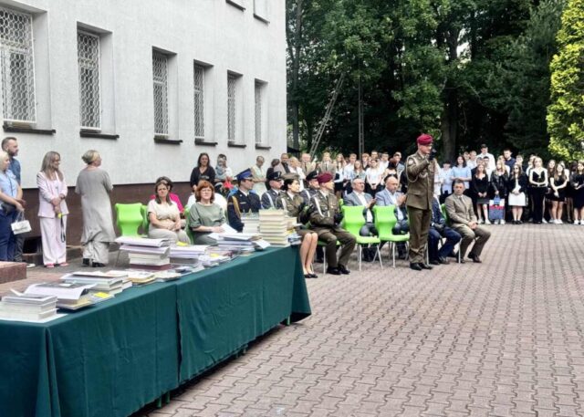 Dziedziniec szkolny, po lewej stronie stoliki przykryte zielonym suknem, na których są nagrody książkowe. Widoczni też nauczyciele, uczniowie i zaproszeni na uroczyste zakończenie roku szkolnego goście.