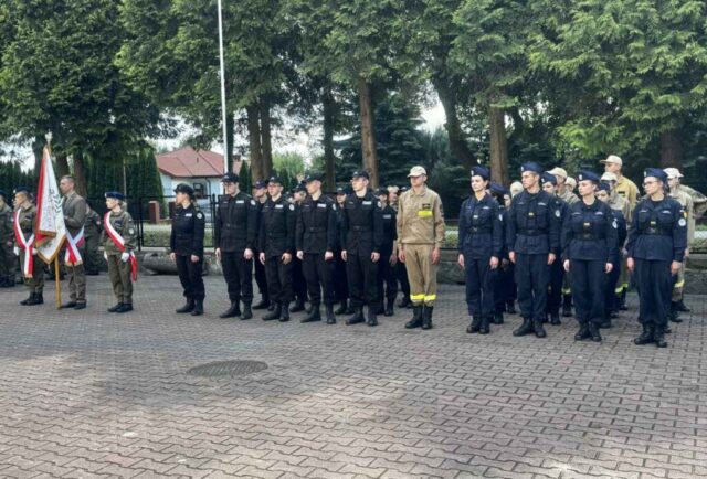 Dziedziniec szkolny, ustawieni w szeregach uczniowie klas mundurowych, po lewej stronie poczet sztandarowy.