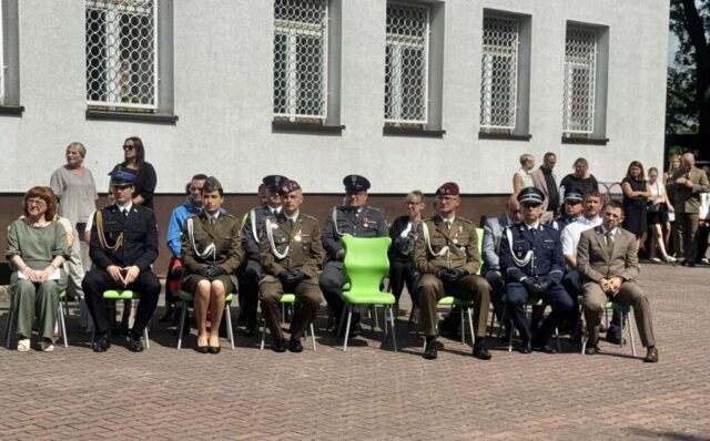 Dziedziniec szkoły, nauczyciele oraz zaproszeni na uroczystość goście. W tle budynek Szkoły.