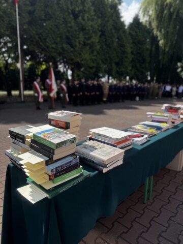 Stoliki przykryte zielonym suknem z widocznymi nagrodami książkowymi. W tle poczet sztandarowy oraz ustawieni w szeregach uczniowie.