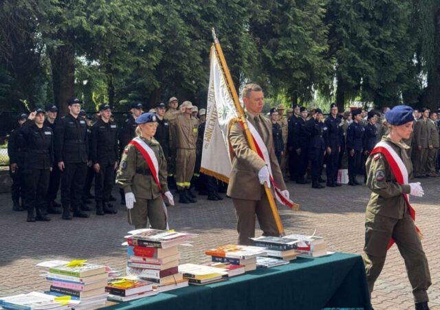 Dziedziniec ZS CKZ w Bujnach, poczet sztandarowy, w tle ustawieni w szeregach uczniowie klas mundurowych i technikum leśnego. W lewym dolnym roku fragment stolików przykrytych zielonym suknem z przygotowanymi do rozdania uczniom nagrodami książkowymi.