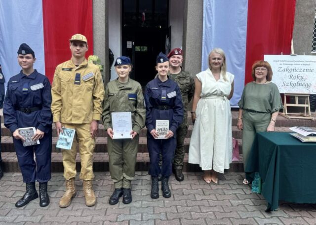 Dyrekcja Szkoły wraz z opiekunem klas OPW i uczniowie klas mundurowych odbierający nagrody książkowe. W tle wejście do budynku Szkoły, flagi Polski i sztaluga z plakatem z napisem Zakończenie Roku Szkolnego.