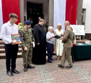 Dziedziniec ZS CKZ Bujny. Dyrekcja, ksiądz i nauczycielka wręczają uczniom nagrody książkowe. Dwóch uczniów w umundurowaniu - klas leśna oraz OPW. W tle flagi Polski i sztaluga z plakatem z napisem Zakończenie Roku Szkolnego. 