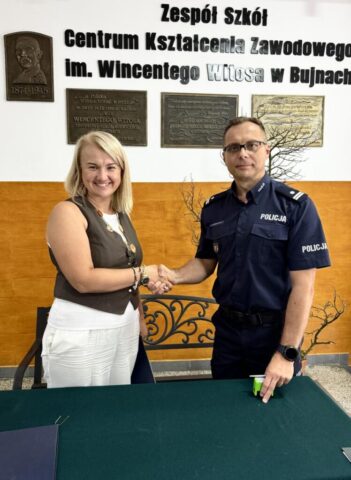 P. Dyrektor ZS CKZ Bujny wraz z komendantem Miejskim Policji w Piotrkowie Tryb stoją i podają sobie ręce. W tle ściana przy wejściu do Szkoły z pełną nazwą szkoły i tablice pamiątkowe.