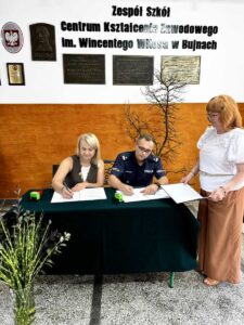 Pani Dyrektor ZS CKZ Bujny oraz Komendant Miejski Policji w Piotrkowie Tryb. siedzą za stołem nakrytym zielonym suknem i podpisują porozumienie. Po prawej stronie stoi p. Wicedyrektor Szkoły. Z przodu, w lewym dolnym rogu widoczny wazon z kwiatami. W tle ściana przy wejściu do Szkoły z pełną nazwą Szkoły oraz tablicami pamiątkowymi. 