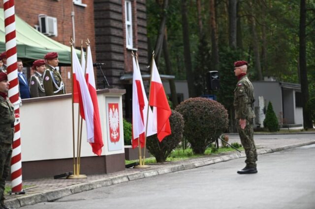 Obchody Święta 25. Batalionu Logistycznego. Po lewej stronie trybuna po prawej stronie żołnierz oddający honory oficjelom. Widoczne też dwa stojaki z Flagami Polski oraz godło Polski.