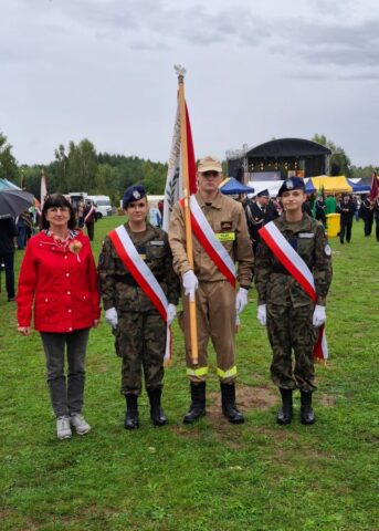 Poczet sztandarowy ZS CKZ Bujny wraz z opiekunem podczas dożynek gminno-powiatowych. W tle inni uczestnicy wydarzenia, stoiska wystawców i scena.