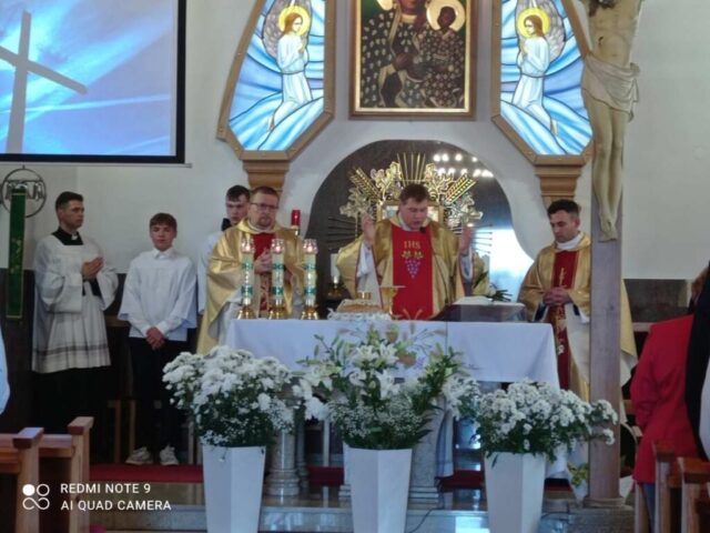 Wnętrze kościoła. 3 księży przy ołtarzu i 3 ministrantów. Dekoracje z białych kwiatów.