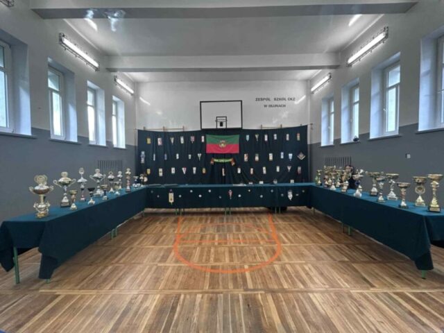 Wnętrze sali gimnastycznej. Ławki ustawione w literę U, przykryte zielonym suknem, na których stoją trofea sportowe. W tle rozwieszone zielone sukno, na którym przywieszone są proporce, szalik i flaga klubu sportowego.