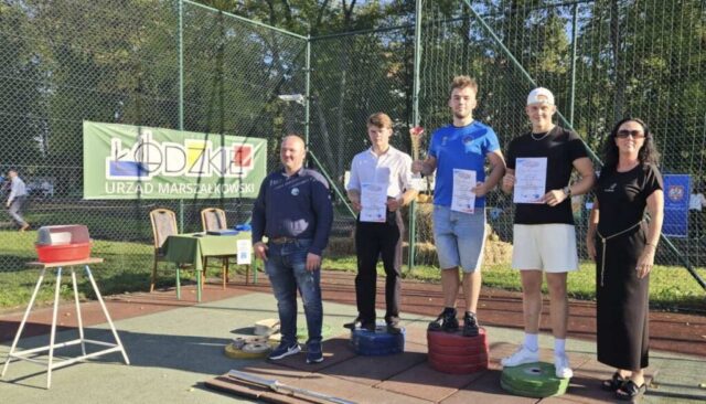 Boisko szkolne orlik. Na podium 3 uczniów, zwycięzców konkursu, po bokach opiekunowie klubu sportowego.