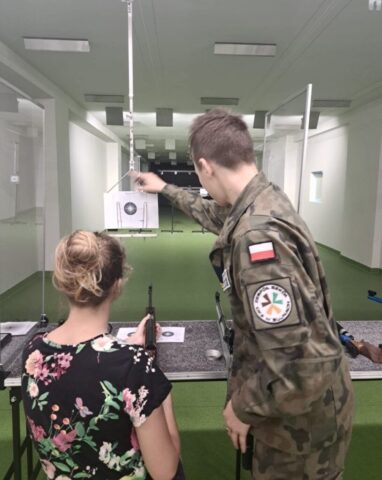 Wnętrze strzelnicy, kadet opw instruuje kobietę, która będzie strzelała do tarczy.