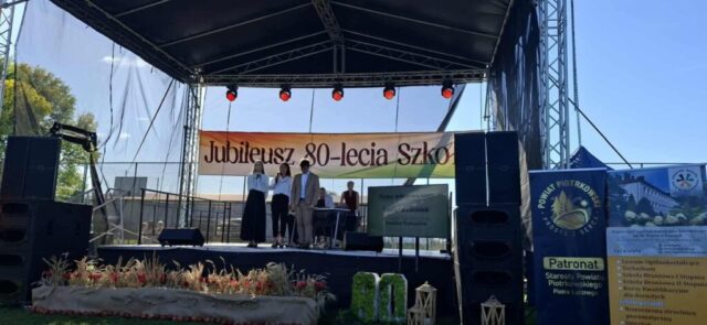 3 osoby na scenie w strojach galowych, w tle dwie dorosłe osoby. Baner z napisem Jubileusz 80-lecia Szkoły. Po prawej stronie rollupy ZS CKZ Bujny oraz Starostwa powiatowego.