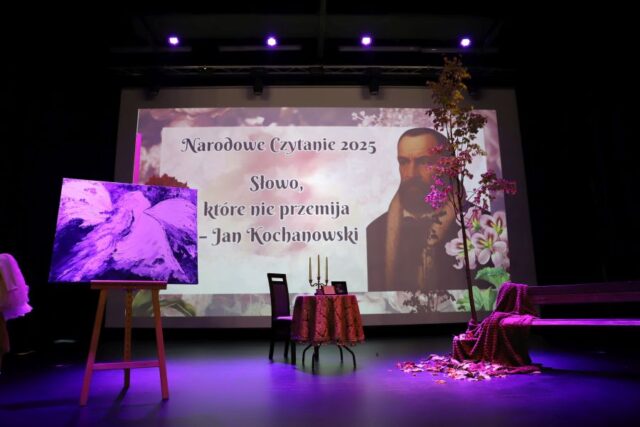 Scena GOK udekorowana na okoliczność Narodowego Czytania. Wyświetlony slajd z cytatem Słowo, które nie przemija i wizerunkiem Jana Kochanowskiego. Na scenie sztaluga z obrazem, stolik i krzesło oraz po prawej stronie ławka.
