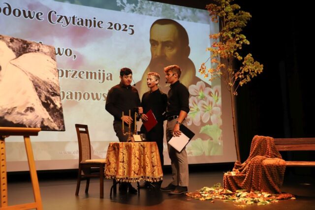 3 uczniów na scenie podczas Narodowego Czytania. Stoją przy stoliku.