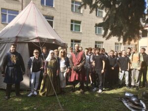 Grupa młodzieży, opiekunowie i osoby w strojach średniowiecznych, przedstawiające rekonstrukcję historyczną. Widoczny także namiot i budynek Akademii Piotrkowskiej. 