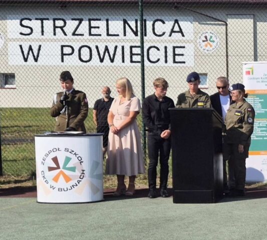 Boisko szkolne Orlik przy ZS CKZ Bujny. Pani Dyrektor, przedstawiciel służb mundurowych, uczniowie klas OPW oraz koordynator OPW. W tle budynek strzelnicy.