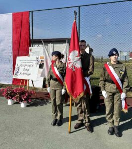 Boisko szkolne orlik, uroczystość rozpoczęcia roku szkolnego. Poczet sztandarowy, w tle również sztaluga z plakatem z napisem rozpoczęcie roku szkolnego, dekoracja z kwiatów w doniczkach oraz zawieszone flagi Polski. 
