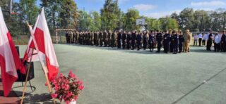 rozpoczecie_roku_szkolnego02 Boisko Szkolne ZS CKZ Bujny, uroczystość rozpoczęcia Roku szkolnego. Uczniowie klas mundurowych oraz inni uczniowie w storjach galowych. Po lewej stronie stojak z flagami Polski oraz dekoracja z kwiatów doniczkowych.