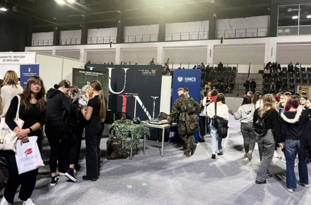Teren wydarzenia Salon Maturzystów, duża hala sportowa, stoiska Uczelni wyższych oraz zwiedzający. W tle trybuny i siedzący na krzesłach uczestnicy Salonu Maturzystów.