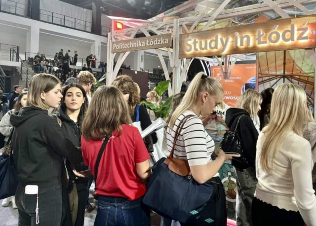 Grupa młodych osób odwiedza stoisko Politechniki Łódzkiej z hasłem Study in Łódź podczas targów edukacyjnych Salon Maturzystów. W tle trybuny i siedzący uczestnicy wydarzenia.
