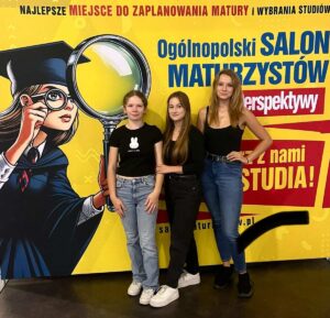 3 uczennice pozują na tle baneru reklamowego wydarzenia pod nazwą Ogólnopolski Salon Maturzystów. Baner zółte tło, po lewej stronie wizerunek uczennicy w todze i birecie, po prawej teksty reklamujące wydarzenie. 