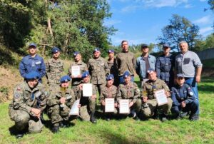 Szkolna drużyna uczniów biorących udział w turnieju KBKS wraz z opiekunami i organizatorami. Pozują do zdjęcia na zewnątrz. W tle drzewa. Większość zawodników w umundurowaniu. 