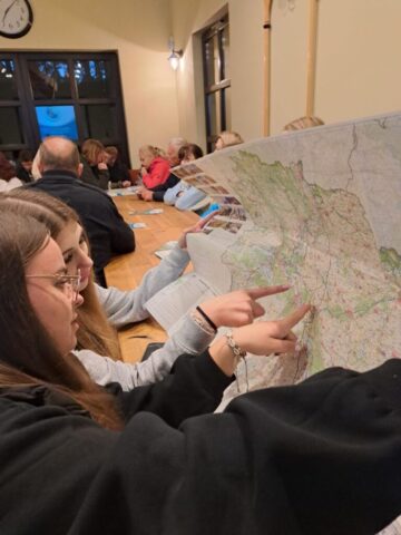 Grupa osób w pomieszczeniu. Siedzą przy stołach. Na pierwszym planie dwie młode kobiety z mapą. Wskazują jakiś punkt na tej mapie.