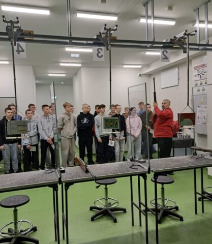 Wnętrze strzelnicy pneumatycznej. Grupa młodzieży przed stanowiskami. Po prawej stronie instruktor.
