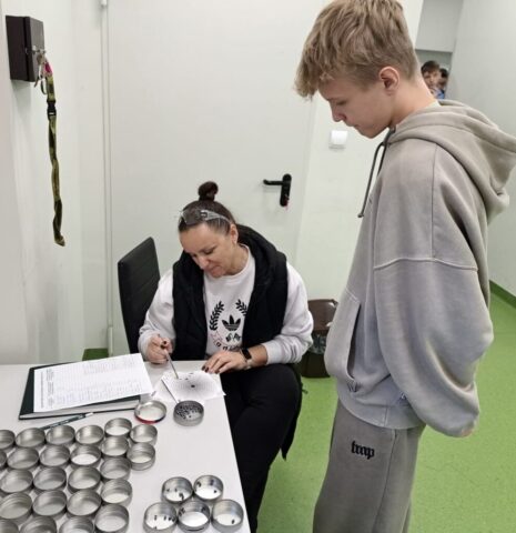 Uczeń przygląda się jak instruktorka wskazuje punkt na papierowej tarczy rozłożonej na biurku. Widoczne też okrągłe, metalowe pojemniki, część z nich zawiera śrut.