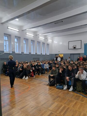 Sala gimnastyczna. Młodzież siedzi na ławkach. Przemawia do nich pani policjantka.